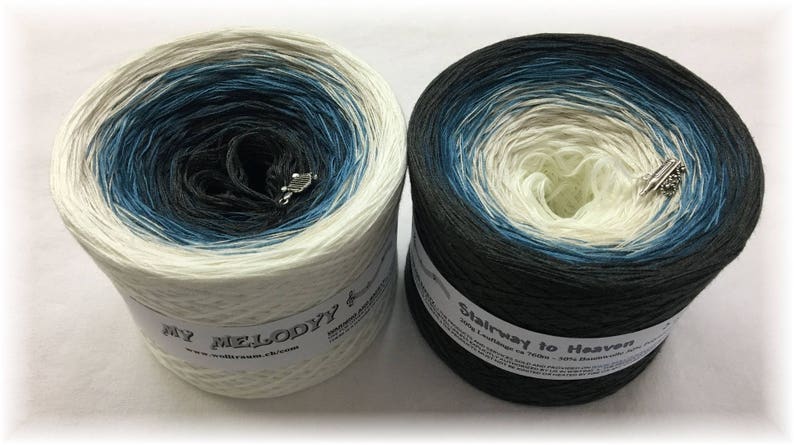 Puede incluir: Dos madejas de hilo con un esquema de color degradado. Una es blanca con azul y negro, la otra es negra con azul y blanco. Cada una tiene una etiqueta y un sujetahilos plateado.
