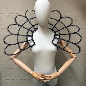 Könnte beinhalten: Ein schwarzes Schulterstück aus Spitze mit offenem, geometrischem Design. Das Stück wird auf einer weißen Schaufensterpuppe mit Holzarmen und -händen präsentiert. Die Spitze hat einen zarten, gewellten Rand.