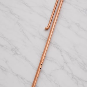May include: A long, slender copper pipe instrument featuring a series of small circular holes along the lower section. The structure includes a curved top section that folds back parallel to the main body, constructed from connected copper tubing.