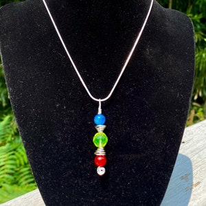 Puede incluir: Un collar de cadena de plata con un colgante que presenta cuatro cuentas en línea vertical. Las cuentas son azules, verdes, verdes y rojas.