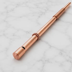 May include: A metallic copper-colored whistle with a cylindrical body, a tapered end, and a decorative twisted rope-like band around the center. The mouthpiece features a circular spiral pattern on the flat end and a small rectangular air vent.