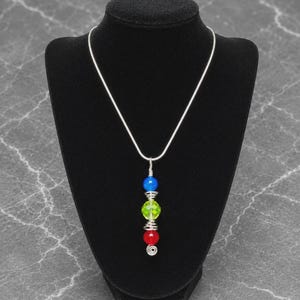May include: A silver-toned snake chain necklace featuring a vertical pendant. The pendant consists of three stacked beads in blue, lime green, and red, separated by silver wire coils. The bottom of the pendant ends in a small silver wire spiral.