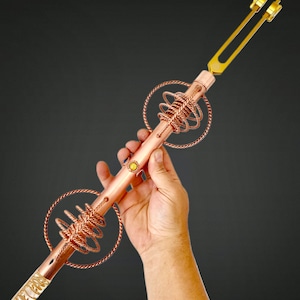 Puede incluir: Un diapasón de cobre con un cristal en la parte inferior y dos anillos de cobre envueltos alrededor del eje. El diapasón se sostiene en una mano.