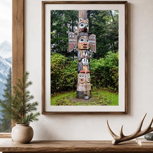 May include: A framed print of a colorful totem pole with carved faces and animal figures. The totem pole is set against a backdrop of lush green trees. The print is displayed on a wooden table with decorative items.