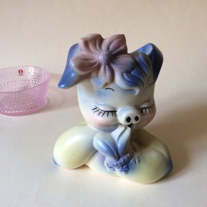 Pode incluir: Uma figura de porquinho de cerâmica com um corpo azul e amarelo, uma flor rosa na cabeça e os olhos fechados. O porquinho está segurando uma flor azul no focinho.