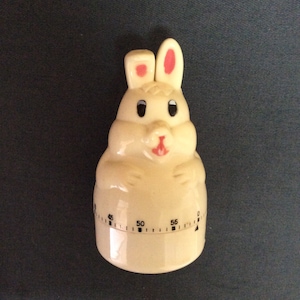 May include: A cream-colored bunny-shaped kitchen timer with black eyes, pink inner ears, and a red nose. The timer has a dial with numbers and tick marks, measuring up to 60 minutes. A cute and functional kitchen accessory.
