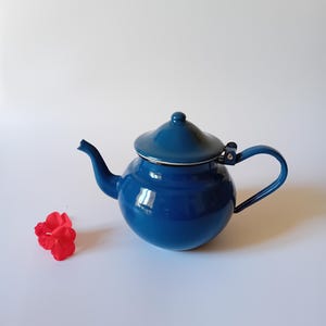 Puede incluir: Una tetera azul cobalto brillante con tapa y asa a juego. La tetera tiene un cuerpo redondeado y un pico curvo. Una pequeña flor roja está a la izquierda de la tetera. La tetera es probablemente para preparar té.