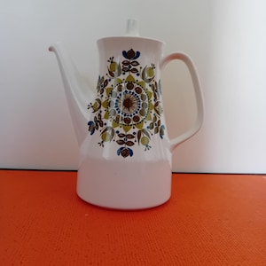 May include: White ceramic coffee pot with a floral pattern in shades of brown, green, and blue. The pot has a long spout, a handle, and a lid. The design is symmetrical and covers the upper portion of the pot.