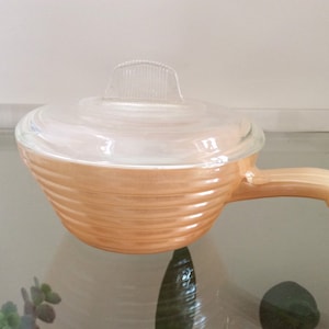 Pode incluir: Um prato de forno de vidro vintage de cor pêssego com uma tampa de vidro transparente e uma alça. O prato tem um design canelado.