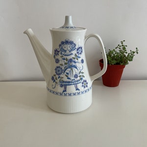 May include: White ceramic coffee pot with a blue and green floral design featuring a stylized woman. The pot has a lid and a handle.