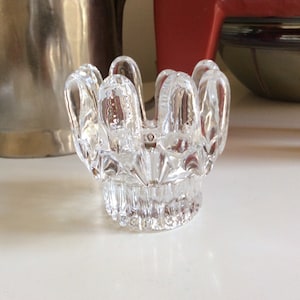 May include: A clear glass candle holder with a decorative, fluted design. The holder has a wide, open top and a narrow base.