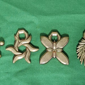 Op de afbeelding: Vijf goudkleurige metalen bedels met verschillende ontwerpen. De bedels zijn in de vorm van een ster, een bloem, een vlinder, een spiraal en een geometrische vorm. De bedels zijn allemaal ongeveer 5 cm lang.