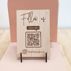 May include: A wooden sign with the text "Follow us" and a QR code. The sign also includes the text "SCAN ME" and a social media icon with the text "littlelaserco".