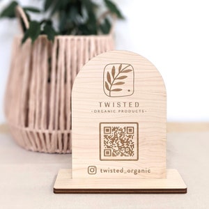 May include: A wooden sign with a rounded top, featuring a stylized leaf logo and the text "TWISTED - ORGANIC PRODUCTS -" along with a QR code and the Instagram handle "twisted_organic".