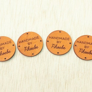 Può includere: Quattro etichette rotonde in pelle marrone chiaro con le parole "HANDMADE BY Phoebe" incise. Ogni etichetta ha quattro piccoli fori sul bordo. Le etichette sono disposte in fila su una superficie di legno chiaro.