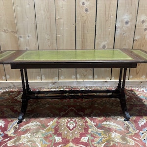 Peut inclure: Une table rectangulaire en bois avec une finition brun foncé et une incrustation en cuir vert sur le dessus. La table comprend deux sections latérales rabattables et repose sur quatre pieds fins et tournés reliés par une entretoise centrale.