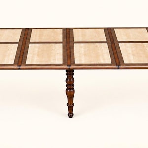 May include: An elongated dining table with a dark brown wooden frame and rounded ends. The tabletop features a series of rectangular, light beige stone inlays arranged in pairs. The table is supported by three turned wooden legs.