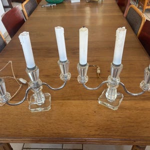 May include: A pair of two-armed candelabras featuring clear glass bases and stems with silver-toned metal arms. Each arm holds a white taper candle. The candelabras are connected by a thin, light-colored electrical cord with a small white switch.