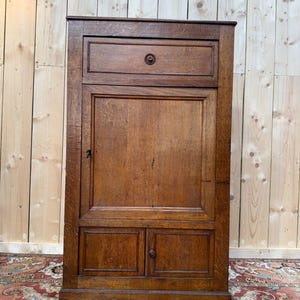 May include: A tall, antique wooden cabinet with a warm, brown finish. It features a drawer at the top with a round knob, a large central door, and two smaller doors at the bottom. The cabinet is likely used for storage.