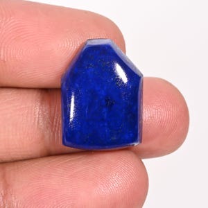 Könnte beinhalten: Ein polierter, tiefblauer Edelstein mit einer einzigartigen, hausähnlichen Form. Der Stein hat eine glatte, reflektierende Oberfläche und eine etwas hellere, fast weiße Oberkante. Der Edelstein wird zwischen Fingern vor weißem Hintergrund gehalten.