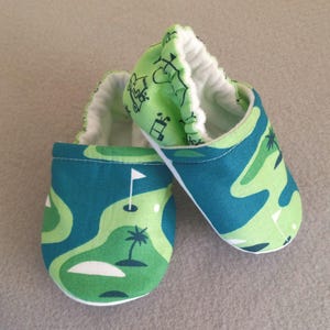 May include: A pair of baby booties with a golf course design. The booties feature a green and blue pattern with images of golf flags, palm trees, and golf carts. The interior is white, and the booties are made of soft fabric.