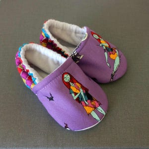 Puede incluir: Un par de zapatillas moradas para beb&eacute; con un dise&ntilde;o de personaje de dibujos animados. Las zapatillas tienen un interior blanco y un ribete estampado en rojo, amarillo y azul. El dise&ntilde;o presenta un patr&oacute;n de patchwork.