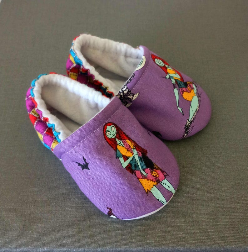 Puede incluir: Un par de zapatillas moradas para beb&eacute; con un dise&ntilde;o de personaje de dibujos animados. Las zapatillas tienen un interior blanco y un ribete estampado en rojo, amarillo y azul. El dise&ntilde;o presenta un patr&oacute;n de patchwork.