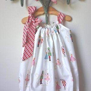May include: A white sleeveless bubble romper featuring a repeating pattern of nutcrackers, ballerinas, and mice. The garment has red and white striped fabric shoulder ties and elasticized leg openings with ruffles, hanging on a wooden hanger.