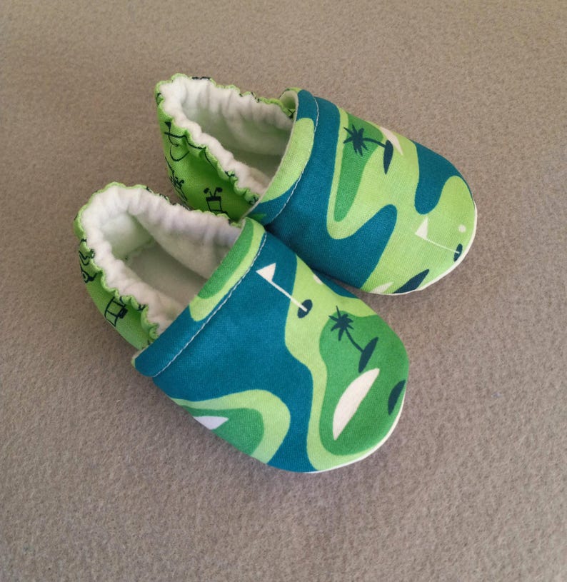 Puede incluir: Un par de zapatos de beb&eacute; con un dise&ntilde;o de campo de golf. Los zapatos presentan un patr&oacute;n de formas verdes y azules con detalles en blanco. El interior es blanco y los bordes est&aacute;n rizados. Zapatos para beb&eacute;s.