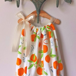 May include: A white baby dress with a pattern of orange fruit and green leaves, hanging on a wooden hanger. The dress has the words "Oh my darling" printed on it. A matching bow is attached to a decorative wall hook.