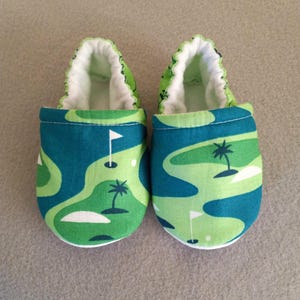 Puede incluir: Un par de zapatos de beb&eacute; con un dise&ntilde;o de campo de golf. Los zapatos presentan un patr&oacute;n verde y azul con banderas de golf y palmeras. El interior es blanco y el ribete es verde.