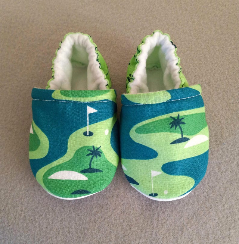 Puede incluir: Un par de zapatos de beb&eacute; con un dise&ntilde;o de campo de golf. Los zapatos presentan un patr&oacute;n verde y azul con banderas de golf y palmeras. El interior es blanco y el ribete es verde.
