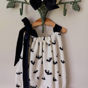 May include: A white romper with a black bat pattern hangs on a wooden hanger. The romper has black ribbon ties and a ruffled hem. A large black bow is attached to a decorative metal hanger.