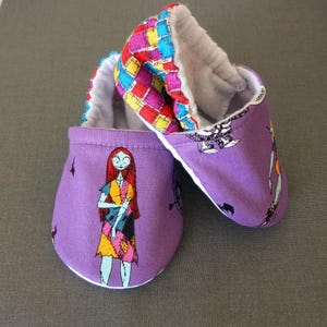 Puede incluir: Un par de patucos de beb&eacute; con exterior morado y dise&ntilde;o de Sally de Pesadilla antes de Navidad. Los pu&ntilde;os tienen un colorido estampado de patchwork y el interior es blanco y suave. Tambi&eacute;n se incluyen peque&ntilde;as formas de murci&eacute;lago negro.