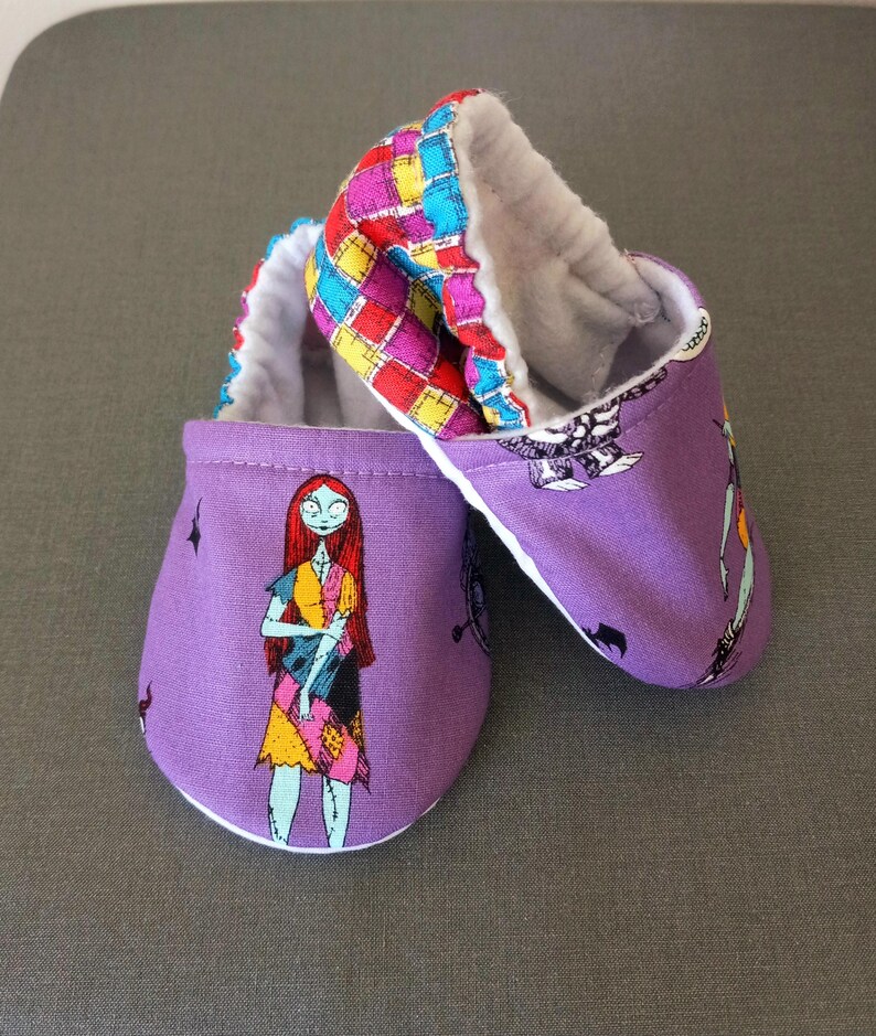 Puede incluir: Un par de patucos de beb&eacute; con exterior morado y dise&ntilde;o de Sally de Pesadilla antes de Navidad. Los pu&ntilde;os tienen un colorido estampado de patchwork y el interior es blanco y suave. Tambi&eacute;n se incluyen peque&ntilde;as formas de murci&eacute;lago negro.