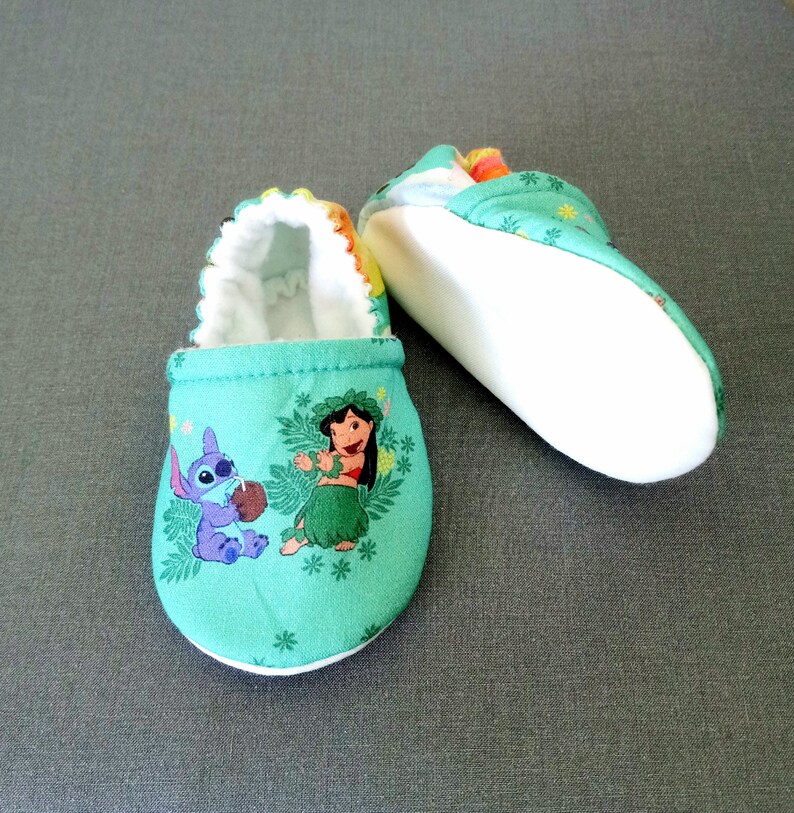 Op de afbeelding: Een paar groene babyschoentjes met een witte voering. De schoentjes hebben een cartoon afbeelding van Lilo en Stitch uit de Disney-film Lilo & Stitch.