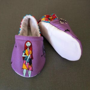 Puede incluir: Un par de patucos morados para beb&eacute; con un dise&ntilde;o de personaje de dibujos animados. Los patucos tienen un interior blanco y un ribete colorido. Un patuco muestra un personaje con pelo rojo y un vestido de retazos. El otro patuco tiene un patr&oacute;n colorido.