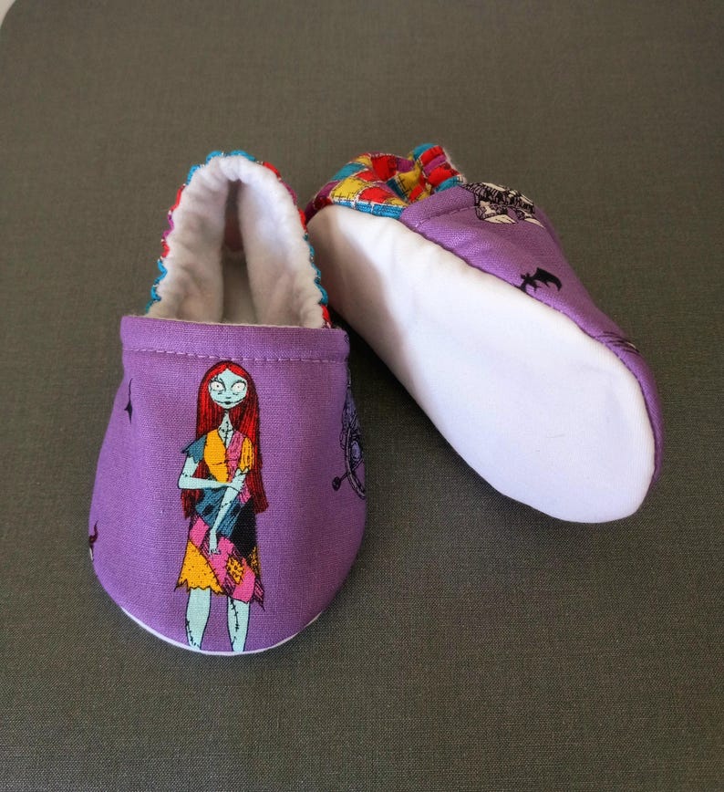 Puede incluir: Un par de patucos morados para beb&eacute; con un dise&ntilde;o de personaje de dibujos animados. Los patucos tienen un interior blanco y un ribete colorido. Un patuco muestra un personaje con pelo rojo y un vestido de retazos. El otro patuco tiene un patr&oacute;n colorido.