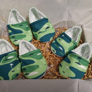 Puede incluir: Seis patucos de beb&eacute; con dise&ntilde;o de campo de golf. Los patucos presentan un estampado verde y azul con forro blanco. Los patucos est&aacute;n colocados en una caja con papel triturado.