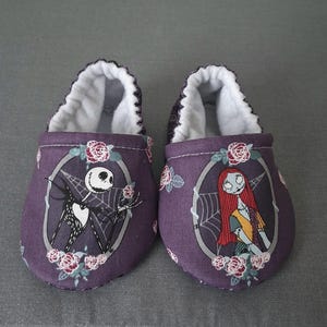 May include: A pair of purple baby shoes featuring Jack Skellington and Sally from The Nightmare Before Christmas. The shoes have a white interior and are decorated with roses. The design is set against a gray background.