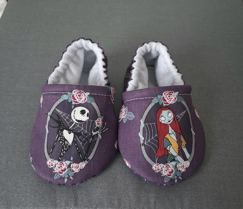 May include: A pair of purple baby shoes featuring Jack Skellington and Sally from The Nightmare Before Christmas. The shoes have a white interior and are decorated with roses. The design is set against a gray background.