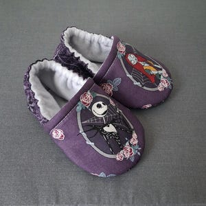 May include: A pair of purple baby booties featuring characters from The Nightmare Before Christmas. Each slipper has a white interior and a decorative image of Jack Skellington and Sally. The booties are decorated with pink roses.