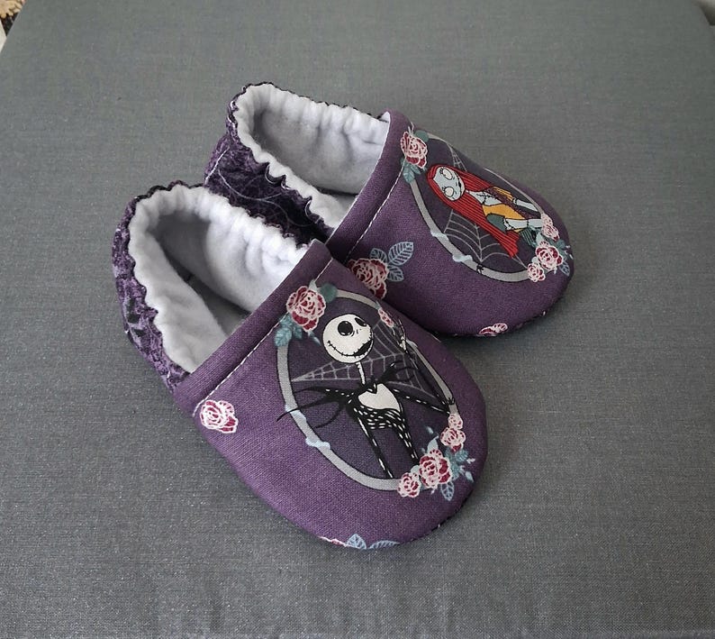 May include: A pair of purple baby booties featuring characters from The Nightmare Before Christmas. Each slipper has a white interior and a decorative image of Jack Skellington and Sally. The booties are decorated with pink roses.
