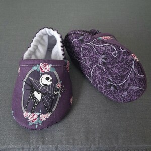 May include: A pair of purple baby booties. One bootie features a Jack Skellington design with roses, while the other has a floral pattern. The interior is white, and the booties are made of soft fabric.