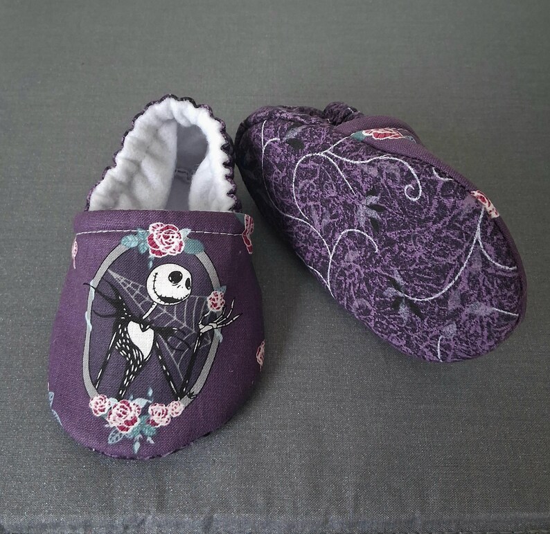 May include: A pair of purple baby booties. One bootie features a Jack Skellington design with roses, while the other has a floral pattern. The interior is white, and the booties are made of soft fabric.