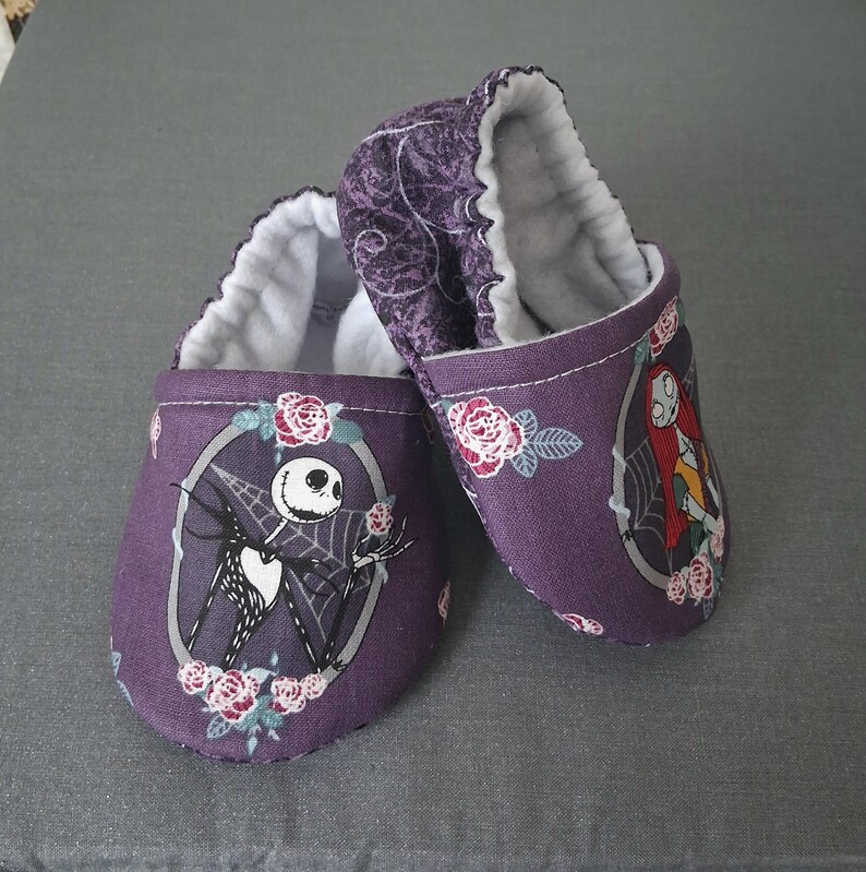 May include: Purple baby booties featuring Jack Skellington and Sally from The Nightmare Before Christmas. The booties have a white interior and are decorated with floral accents. The design is set against a gray background.
