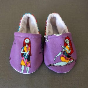 Puede incluir: Un par de patucos morados para beb&eacute; con una colorida ilustraci&oacute;n de Sally de Pesadilla antes de Navidad. Los patucos tienen un interior blanco y un ribete estampado.