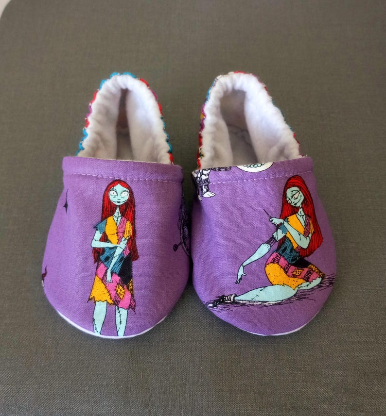 Puede incluir: Un par de patucos morados para beb&eacute; con una colorida ilustraci&oacute;n de Sally de Pesadilla antes de Navidad. Los patucos tienen un interior blanco y un ribete estampado.