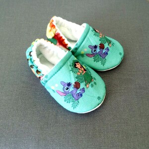 Op de afbeelding: Een paar baby-slofjes in tealblauw met een witte rand. De slofjes hebben een cartoonontwerp van Stitch en Lilo uit de Disney-film Lilo & Stitch.