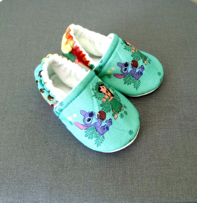 Op de afbeelding: Een paar baby-slofjes in tealblauw met een witte rand. De slofjes hebben een cartoonontwerp van Stitch en Lilo uit de Disney-film Lilo & Stitch.